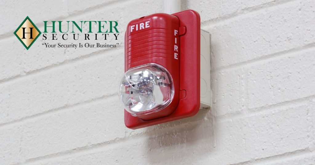 Red commercial fire alarm strobe light mounted on white brick wall with Hunter Security logo