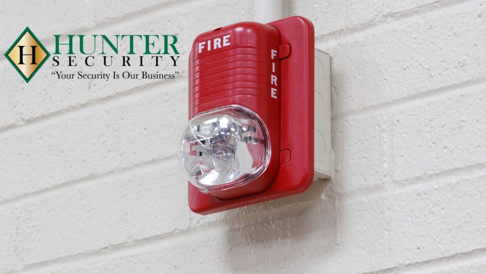 Red commercial fire alarm strobe light mounted on white brick wall with Hunter Security logo